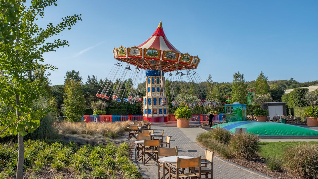 Vibrant carousel in amusement park with tables in foreground and lush greenery.